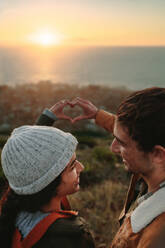 Nahaufnahme eines jungen Mannes und einer Frau, die zusammen auf einem Berggipfel stehen und ein Herz mit den Händen gegen den Sonnenuntergang bilden. Verliebtes Paar, das mit den Fingern ein Herz bildet. - JLPSF21260