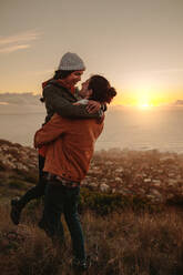 Romantic couple standing on mountain peak at sunset. Man carrying his girlfriend in his arms on cliff. - JLPSF21261