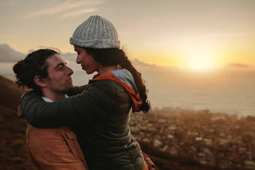 Romantic couple hugging and looking at each other with love. Man carrying woman on winter day. Both on mountain peak during sunset. - JLPSF21262