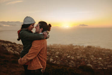 Loving couple standing on mountain peak at sunset. Man carrying his woman in his arms on cliff with seascape in background. - JLPSF21263