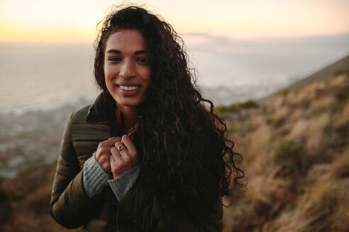 Porträt einer jungen Afrikanerin mit lockigem Haar und warmer Kleidung, die auf einer Klippe steht, eine Frau in der Natur an einem Wintertag. - JLPSF21264