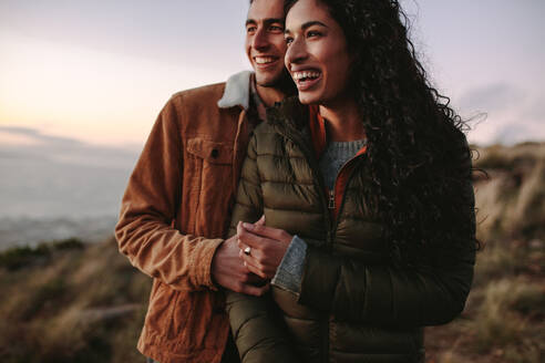 Traveling couple in winter wear standing outdoors together on cliff and looking at view. Man and woman looking at view from mountain peak and laughing. - JLPSF21267