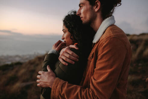 Romantisches junges Paar, das zusammen in den Bergen steht und die Aussicht genießt. Junger Mann umarmt seine Freundin und schaut weg. - JLPSF21271