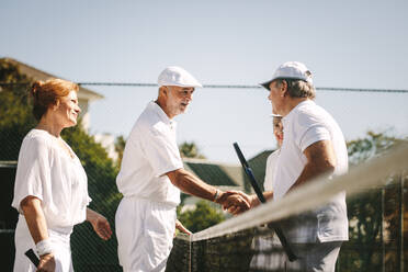 Ältere Männer, die sich nach dem Spiel auf dem Tennisplatz die Hände schütteln. Senioren, die sich nach einem Spiel des gemischten Doppel-Tennis am Netz stehend begrüßen. - JLPSF21279