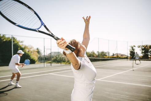 Rückansicht einer Frau, die den Ball bei einem gemischten Doppel aufschlägt. Männer und Frauen spielen an einem sonnigen Tag Tennis im Freien. - JLPSF21280