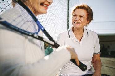 Ältere Frauen sitzen auf einer Bank in der Nähe eines Tennisplatzes und unterhalten sich. Lächelnde Frauen in Tenniskleidung, die sich während eines Tennisspiels ausruhen. - JLPSF21282