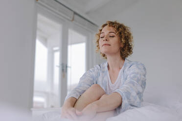 Frau sitzt auf dem Bett mit geschlossenen Augen nach dem Aufwachen am Morgen. Frau sitzt friedlich auf dem Bett mit geschlossenen Augen. - JLPSF21294