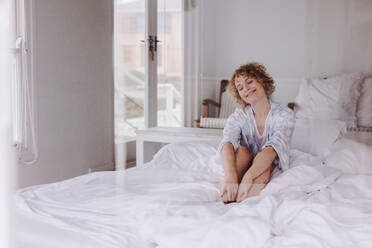 Happy woman sitting on bed after waking up in the morning. Woman waking up in the morning with a smile sitting on bed in her bedroom. - JLPSF21295