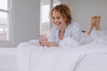 Happy woman lying on bed checking her cell phone. Woman using her mobile phone lying in bed after waking up in the morning. - JLPSF21296