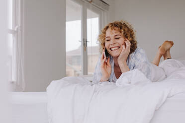 Lächelnde Frau, die nach dem morgendlichen Aufwachen im Bett liegt und mit ihrem Handy spricht. - JLPSF21297