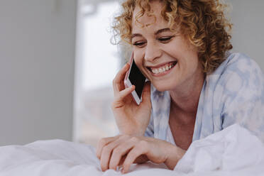 Lächelnde Frau, die auf dem Bett liegend mit einem Handy spricht. Frau in entspannter Stimmung, die mit einem Handy spricht. - JLPSF21299