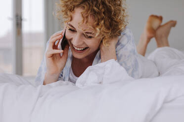 Frau spricht mit Handy während sie sich auf dem Bett entspannt. Frau spricht mit Handy im Bett liegend. - JLPSF21300