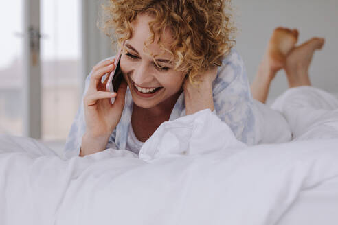 Frau spricht mit Handy während sie sich auf dem Bett entspannt. Frau spricht mit Handy im Bett liegend. - JLPSF21300
