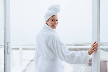 Lächelnde Frau im Bademantel mit einem Handtuch um den Kopf gewickelt. Rückansicht einer Frau, die nach ihrem Bad auf dem Balkon steht. - JLPSF21302