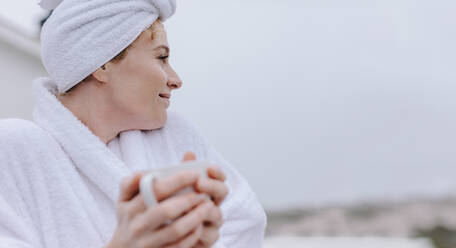 Woman in bathrobe sitting in balcony holding a coffee cup. Woman drinking coffee enjoying the morning weather. - JLPSF21303