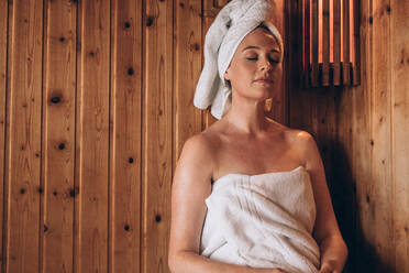 Frau mit Handtuch um ihren Körper und Kopf gewickelt sitzt in der Sauna Spa friedlich. Frau entspannt in einem hölzernen Spa mit geschlossenen Augen. - JLPSF21306