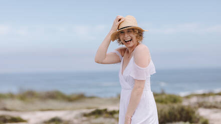 Lächelnde Frau mit Hut vor dem Meer im Hintergrund. Fröhliche Frau in weißem Kleid, die lachend ihren Hut hält. - JLPSF21312