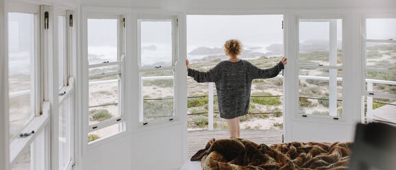 Frau steht an der Balkontür eines Strandhauses und genießt den Blick aufs Meer. Frau in dickem Pullover schaut aufs Meer. - JLPSF21315