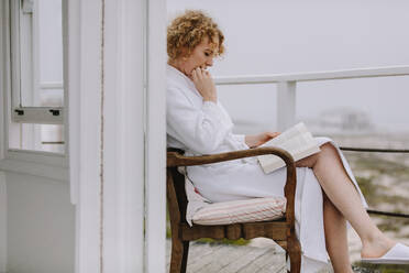 Frau sitzt auf dem Balkon eines Strandhauses und liest ein Buch. Frau entspannt sich beim Lesen eines Buches auf einem Stuhl sitzend. - JLPSF21321