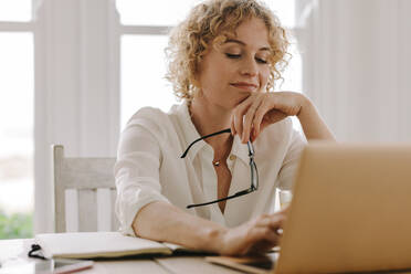 Woman working from home with laptop and diary on the table. Woman holding her eyeglasses in hand and using laptop. - JLPSF21325