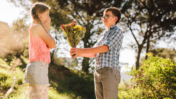 Seitenansicht eines Jungen, der seiner Freundin in einem Park einen Heiratsantrag mit Blumen macht. Ein Junge, der einem Mädchen in einem Park Blumen schenkt, wobei das Mädchen seine Überraschung zum Ausdruck bringt. - JLPSF21340