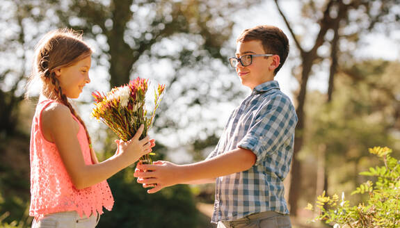 Junge schenkt einem Mädchen in einem Park Blumen. Seitenansicht von verliebten Kindern in einem Park mit einem Blumenstrauß. - JLPSF21341