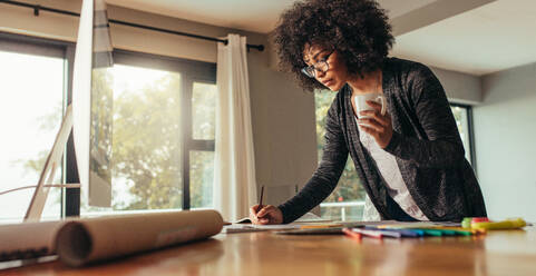 Architect working on plans and blueprints. Woman with cup of coffee making notes in a book at home office. - JLPSF21347