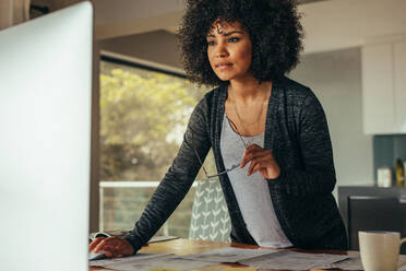 Weibliche Innenarchitektin mit Brille in der Hand arbeitet am Computer im Home Office. Junge afrikanische Frau steht an ihrem Schreibtisch und schaut auf den Computerbildschirm. - JLPSF21348