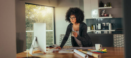 Woman interior designer with color swatch in hand working on computer in the home office. Young african woman standing at the table and looking for a color while making design on computer. - JLPSF21349