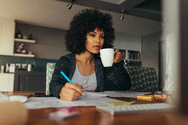 Afrikanische Frau, die an ihrem Schreibtisch arbeitet, Notizen macht, auf den Computerbildschirm schaut und Kaffee trinkt. Architektin, die an ihrem Schreibtisch im Heimbüro arbeitet. - JLPSF21355