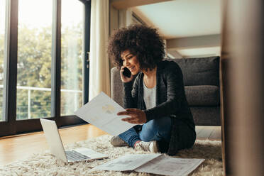 Smiling young woman sitting on floor with few reports talking on cell phone. African female working from home. - JLPSF21365