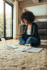 Business woman sitting on floor in living room with laptop and reports lying around texting on mobile phone. African female with curly hair using phone while sitting on floor at home. - JLPSF21366