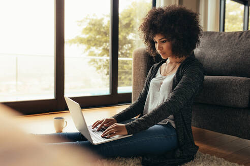 Junge Frau sitzt auf dem Boden neben der Couch und benutzt einen Laptop. Afrikanische Frau arbeitet zu Hause an einem Laptop Computer. - JLPSF21367