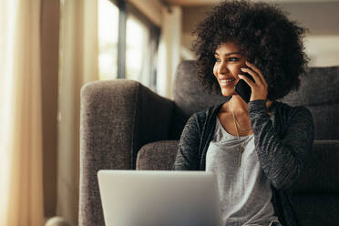 Schöne junge Frau zu Hause mit Laptop und sprechen auf Handy und lächelnd. Weibliche mit Handy Handy, während zu Hause sitzen. - JLPSF21369