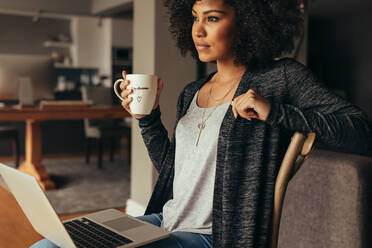 Young african woman sitting in living room with laptop and cup of coffee looking away thinking. Thoughtful female having coffee while working on laptop at home. - JLPSF21373