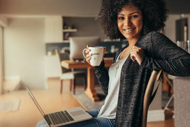 Porträt einer schönen jungen Frau, die zu Hause mit einem Laptop sitzt und Kaffee trinkt. Afrikanische Frau, die sich zu Hause mit Kaffee und Laptop entspannt, in die Kamera schaut und lächelt. - JLPSF21374