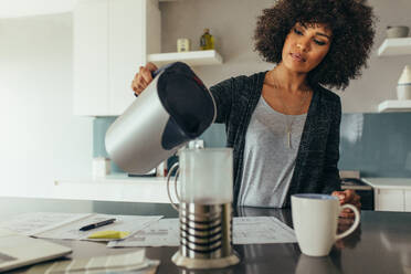 Junge Frau sitzt an ihrem Arbeitstisch und gießt Wasser in einen Krug. Afrikanische Frau macht Kaffee für sich selbst, während sie im Büro zu Hause arbeitet. - JLPSF21379
