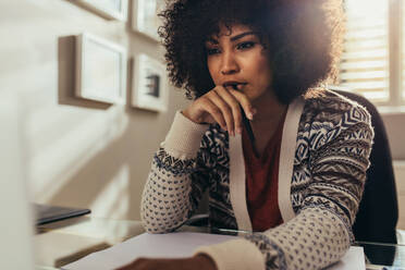 Nachdenkliche junge Frau, die wegschaut, während sie einen Bauplan am Tisch im Büro zu Hause zeichnet. Afrikanische Frau arbeitet an einem neuen Grundriss im Büro und denkt nach. - JLPSF21399