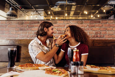 Junger Mann und Frau sitzen in einem Fast-Food-Restaurant und füttern sich gegenseitig mit Pizza. Paar genießt Pizza im Café. - JLPSF21400
