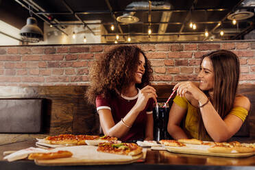 Zwei junge Frauen mit Pizza und Cola auf einem Tisch in einem Fastfood-Restaurant. Freundinnen in einem Restaurant, die sich gegenseitig ansehen und ein kaltes Getränk mit zwei Strohhalmen teilen. - JLPSF21401