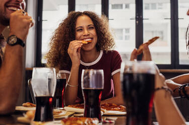 Lächelnde junge Frau mit lockigem Haar isst Pizza mit Freunden im Café. Frau mit Freunden genießen Pizza mit Essen und Getränken in Tabelle. - JLPSF21402