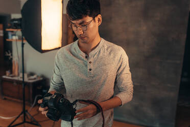 Asiatischer männlicher Fotograf, der seine DSLR-Kamera im Studio überprüft und einstellt. Fotograf, der seine Kamera vor der Aufnahme im Studio einstellt. - JLPSF21403
