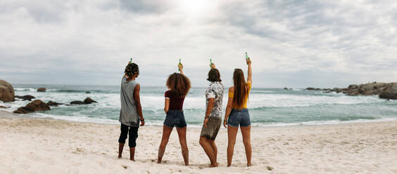 Rückansicht von jungen Freunden, die am Strand stehen und eine Bierflasche ins Meer halten. Eine Gruppe von Freunden feiert und trinkt am Strand. - JLPSF21408