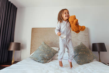 Fröhliches kleines Mädchen springt auf dem Bett mit Teddybär. Kind Mädchen spielt auf dem Bett in ihrem Schlafzimmer und lächelnd. - JLPSF21419
