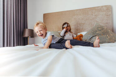 Glückliche Kinder spielen im Schlafzimmer. Kleiner Junge lächelnd mit Mädchen auf der Rückseite in Krone auf dem Bett. - JLPSF21422