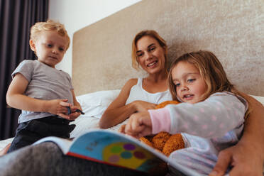 Junge Mutter mit ihren Kindern auf dem Bett, die ein Märchenbuch lesen. Niedliches kleines Mädchen, das auf ein Märchenbuch zeigt, während sie mit ihrer Mutter und ihrem Bruder auf dem Bett sitzt. - JLPSF21424