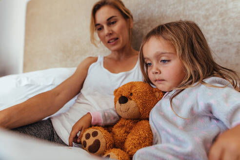 Kleines Mädchen liest ein interessantes Buch auf dem Bett sitzend mit ihrer Mutter. Mutter und Tochter lesen ein Buch im Bett. - JLPSF21425