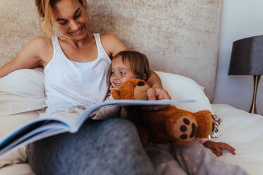 Happy family reading bedtime story in bed. Woman looking at her daughter and smiling while reading a book in bedroom. - JLPSF21427