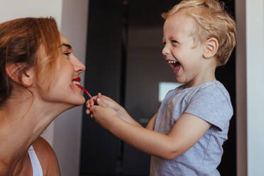 Smiling little boy putting on the makeup to her mother. Happy mother and son playing with makeup at home. - JLPSF21433