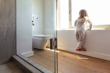 Kleines Mädchen im Badezimmer, das an einem großen Fenster steht und nach draußen schaut. Mädchenkind im Badezimmer, das durch ein Fenster nach draußen schaut. - JLPSF21442